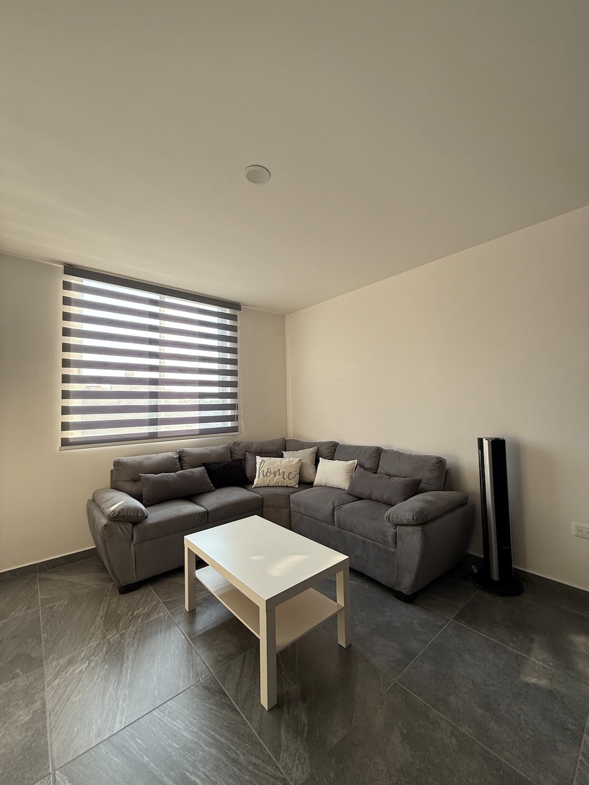 A modern living area features a comfortable gray sectional sofa arranged around a small, white coffee table. Natural light enters through large, horizontal striped windows, illuminating the clean lines of the minimalist design. A slim black tower fan is positioned nearby, providing efficient air circulation.