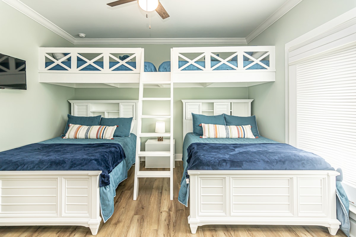 A bright bunk room features two large beds with white frames, accompanied by a lofted bunk arrangement above. Soft blue bedding and decorative pillows enhance the sleeping areas. A simple ladder provides access to the upper bunks, while a wall-mounted TV adds to the room's functionality.