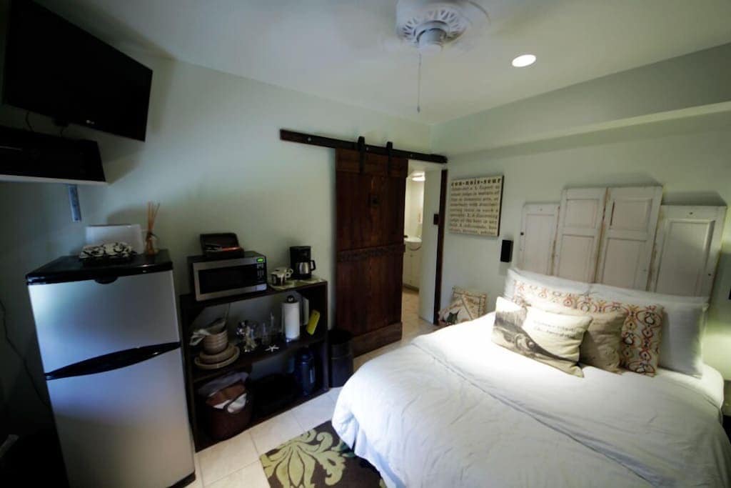A comfortable studio space features a queen bed adorned with decorative pillows. A small fridge, microwave, and coffeemaker are conveniently arranged on a shelf. A wall-mounted TV is visible above, and sliding barn doors lead to the bathroom area, enhancing the room's functionality.