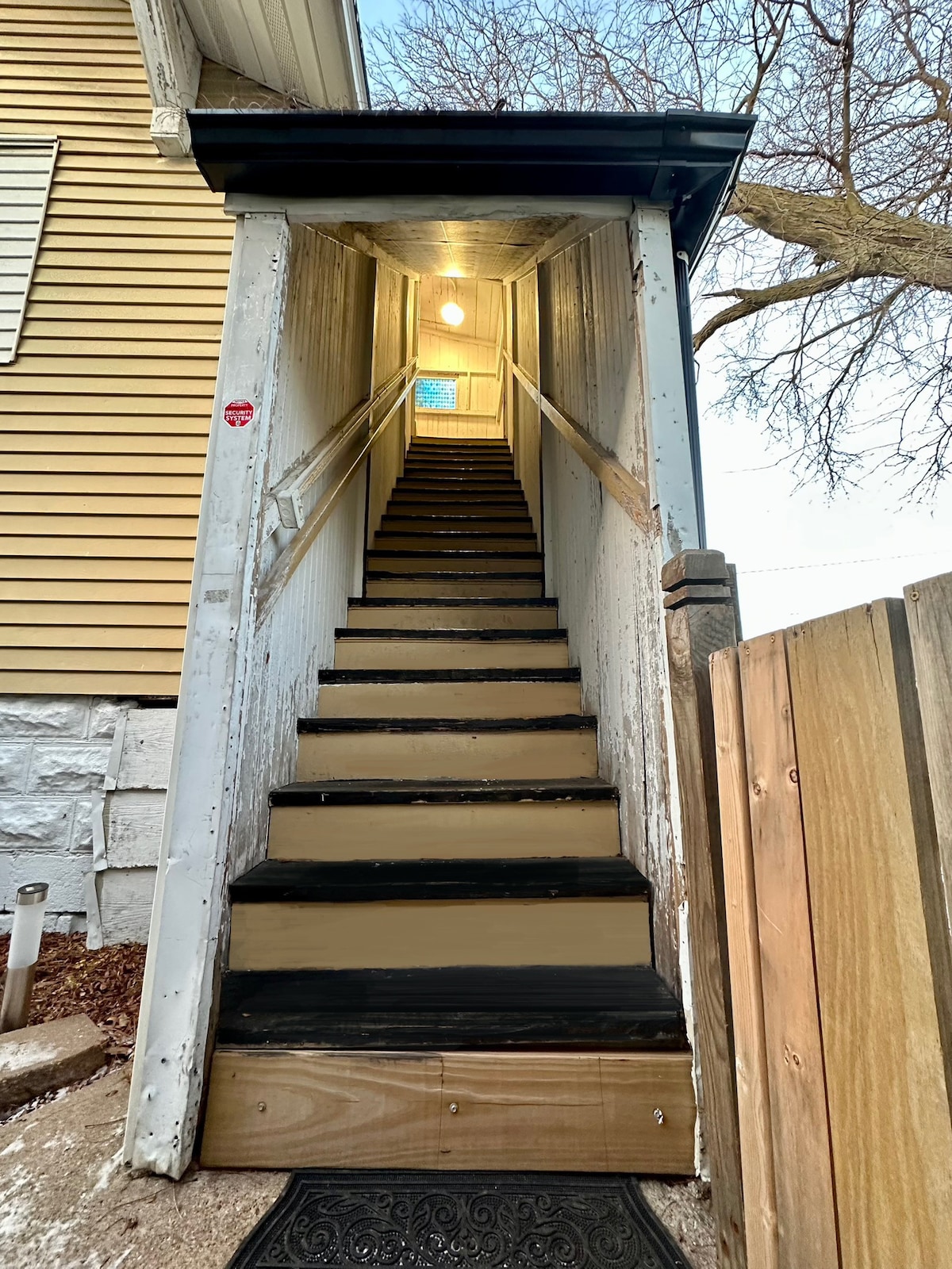 The external stairs lead to a private entrance, framed by walls that are partially stained. A light fixture illuminates the stairway, which features a straight ascent to the upper level. The trees and sky are visible at the top of the stairs.