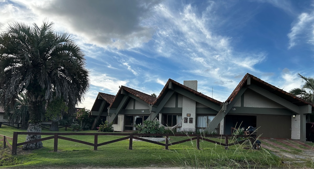 The exterior of a single-story house is depicted, featuring a classic beach house design with a sloped roof and natural wood accents. Lush greenery surrounds the property, with palm trees visible. A wooden fence outlines the yard, and a two-car garage is seen on the right.