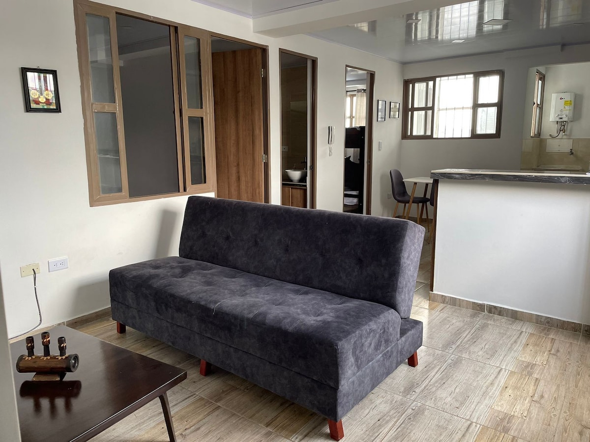 A spacious living area features a dark gray sofa positioned in front of a wooden table. Natural light enters through large windows, illuminating the neutral walls. The kitchen area is partially visible in the background, showcasing a modern design.