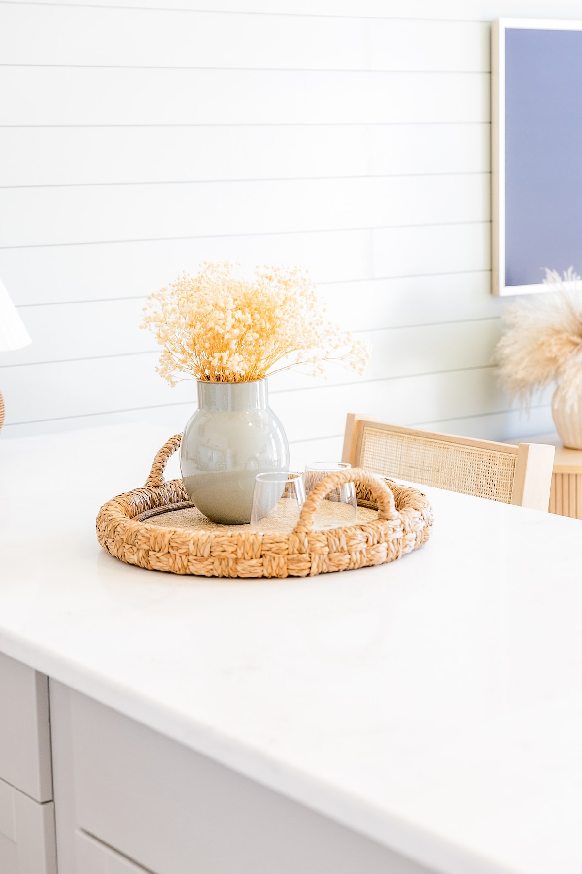 A modern kitchen countertop is highlighted by a woven tray featuring a soothing vase filled with dried flowers. Two clear glasses sit next to the vase. Soft shiplap walls and a muted wall art piece create a subtle backdrop.