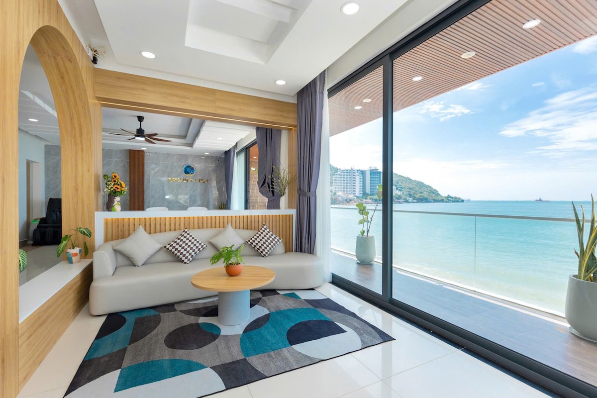 A modern living area features expansive glass doors that open to stunning ocean views. A light-colored sofa with decorative cushions is positioned around a circular coffee table. A vibrant area rug adds a touch of colour to the space, complemented by natural light streaming in.