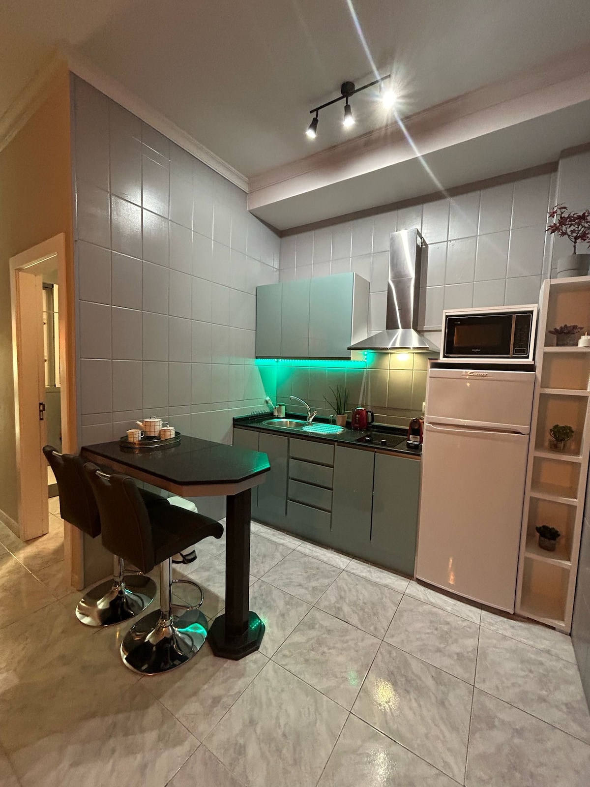 A compact kitchen space is presented, featuring sleek gray cabinetry and modern appliances. A white microwave and silver refrigerator are visible. The area is illuminated by overhead lights, and two high stools are positioned at a small table, creating a cozy dining nook.