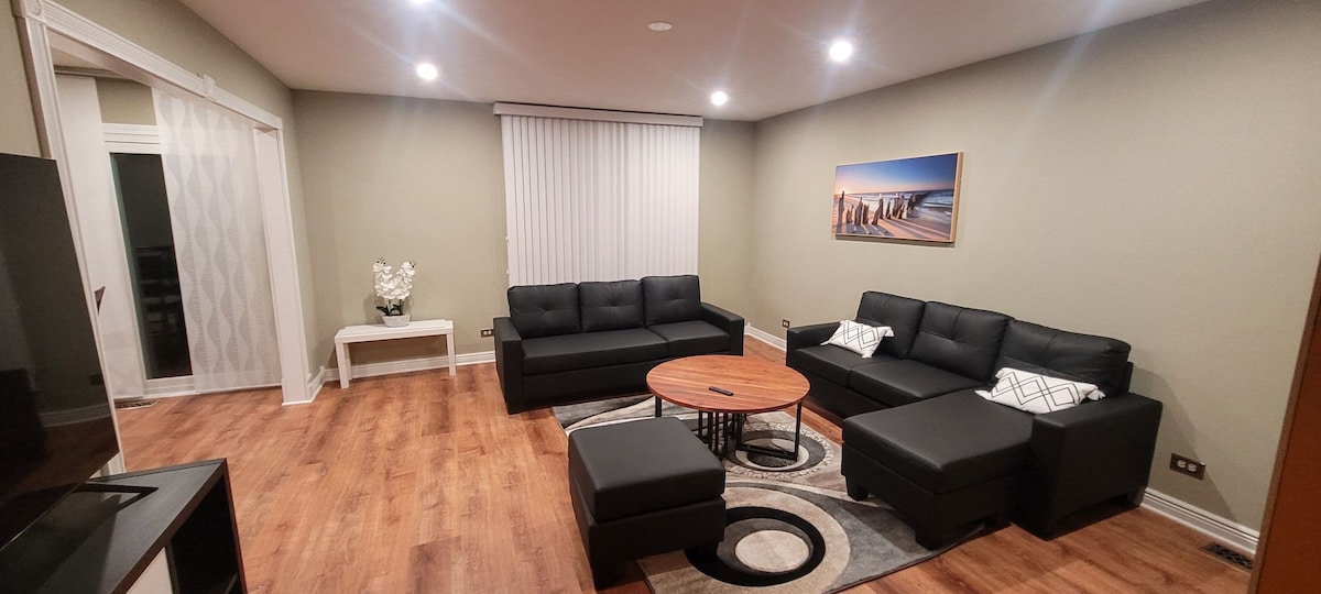 A well-arranged living room is illuminated by recessed lighting, featuring black sofas positioned around a circular coffee table. A decorative area rug complements the furnishings, while a piece of wall art depicting a beach scene adds interest. Large windows are covered with vertical blinds.