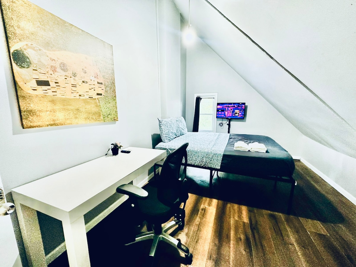 A spacious bedroom is showcased, featuring a cozy bed covered with a patterned blanket. A desk with an ergonomic chair is positioned beside the bed, providing a workspace. A wall-mounted smart TV is visible, along with a piece of art above the desk and sloped ceilings.