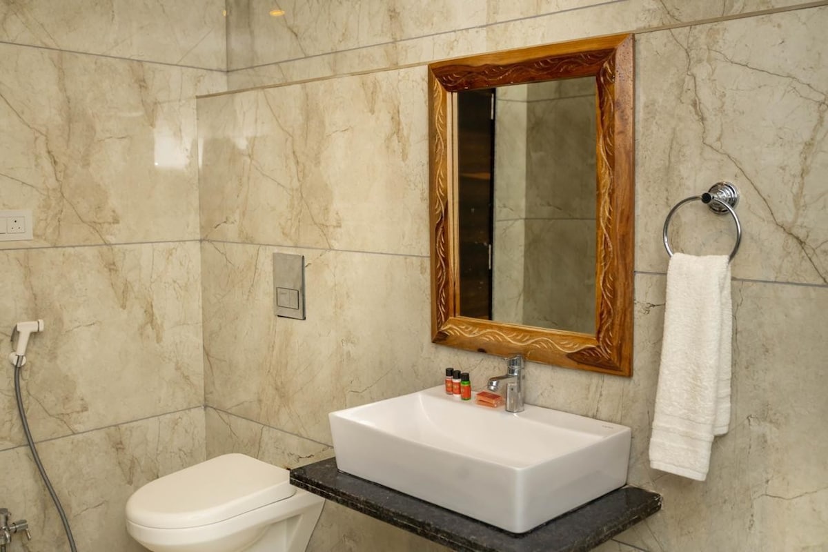 A modern bathroom features neutral-toned marble walls and a sleek countertop. A rectangular sink is positioned on a dark surface, complemented by a decorative wooden mirror above. Toiletries are arranged neatly nearby, and a white towel hangs on a chrome ring, creating a functional space.