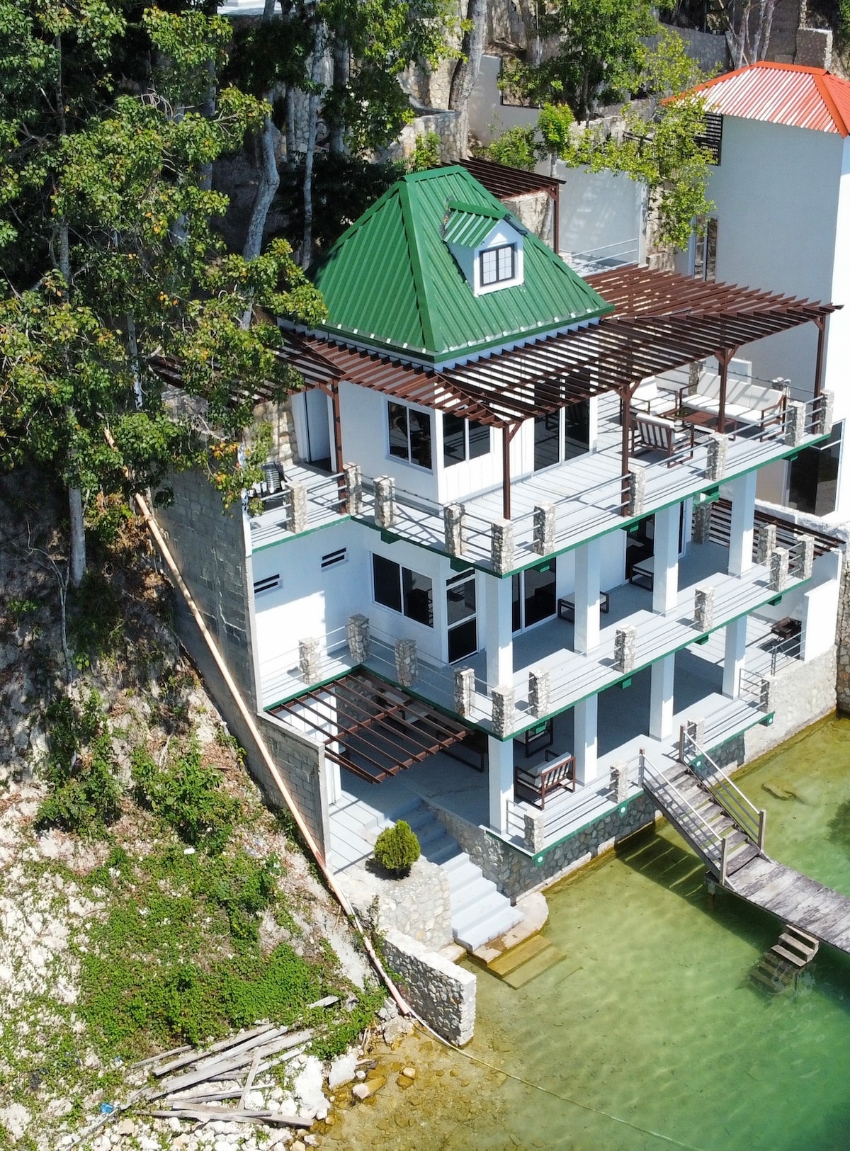 A multi-level property is situated along the water's edge, featuring a green roof and spacious balconies on each level. Surrounding trees add natural beauty, while a wooden dock extends towards the calm water, enhancing accessibility to the lakeside.
