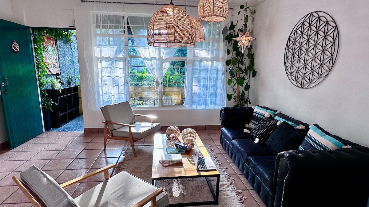 A bright living area features large windows dressed in sheer curtains, allowing natural light to illuminate the space. Woven lamps hang from the ceiling, providing soft overhead lighting. A comfortable sofa, accented with cushions, faces a wooden coffee table, while two armchairs add additional seating.