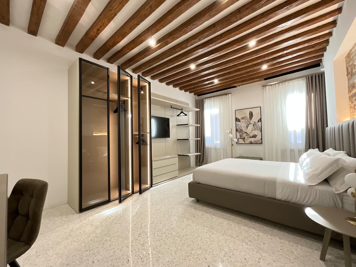 A spacious bedroom is highlighted by wooden beams on the ceiling, complemented by light-colored walls. A large closet with glass doors and a wall-mounted TV offer modern amenities, while soft bedding and a minimalistic design enhance the room's comfort. Natural light filters in through sheer curtains.