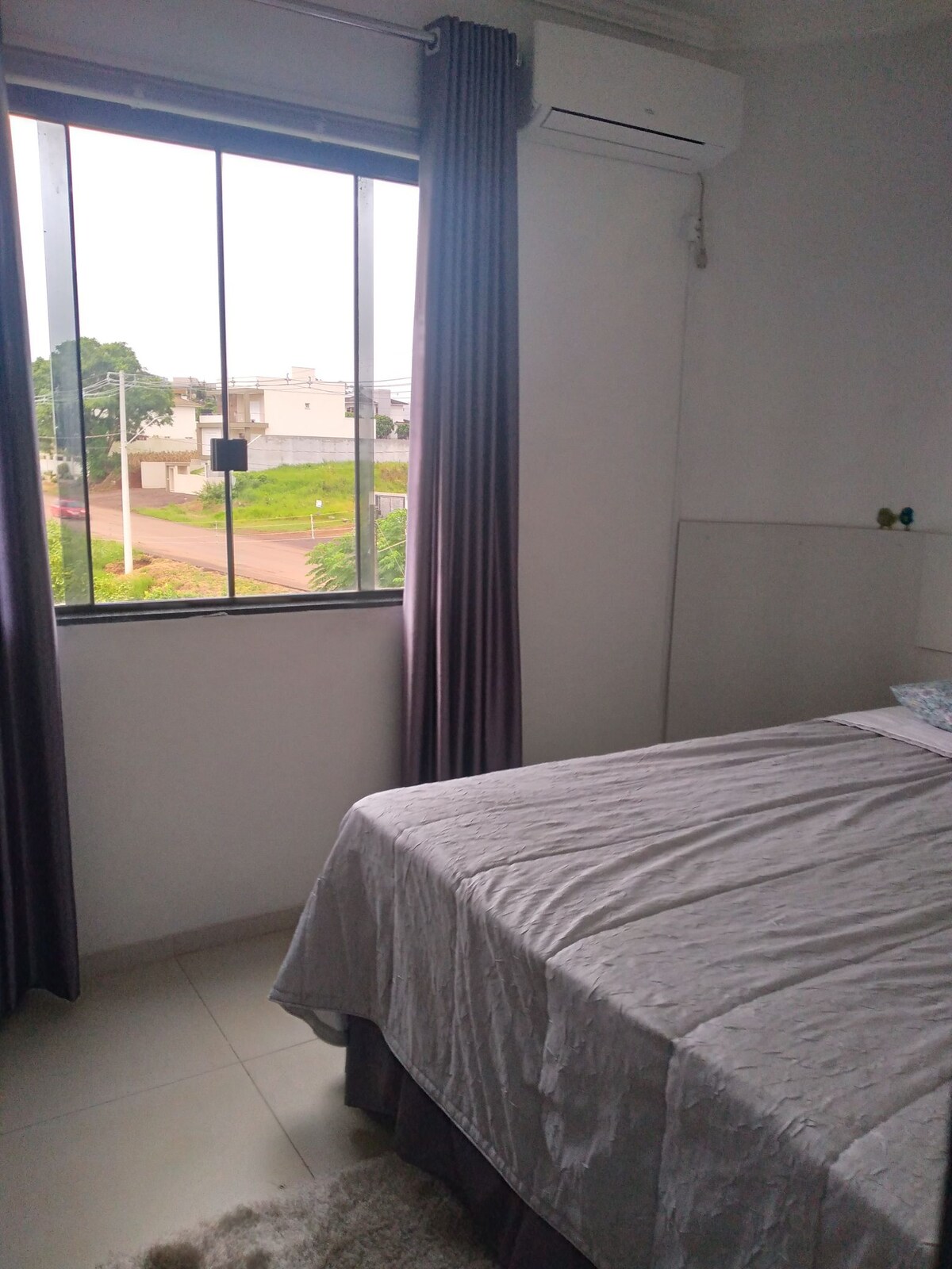 A bright sleeping area features a spacious bed covered with a light grey blanket. Sheer curtains frame a large window, providing views of the greenery outside and natural light. A minimalist design is complemented by a wall mirror and a small decorative item on the ledge.