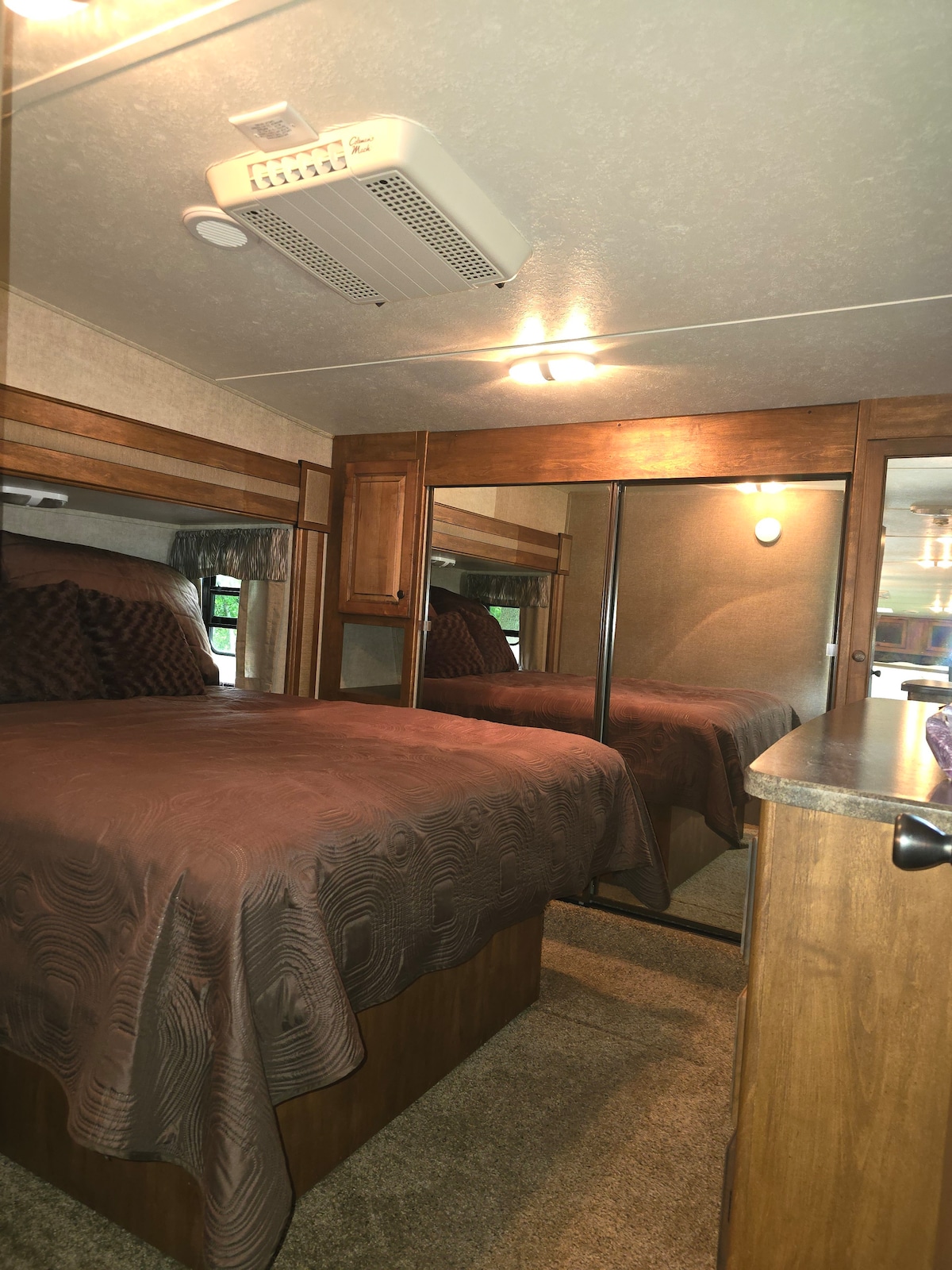 A cozy sleeping area is featured with a brown bedspread draped over a spacious bed. Mirrored closet doors reflect the warm wood accents and soft lighting above. The carpeted floor enhances the comfortable ambiance of the room.