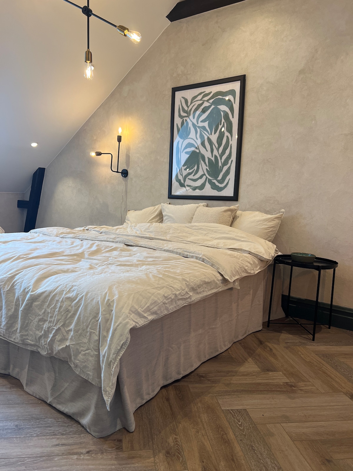 A comfortable sleeping area features a spacious bed dressed in light, soft linens. A framed botanical artwork is displayed above the bed, complemented by a wall-mounted sconce. A small nightstand sits beside the bed, offering a subtle touch of style.