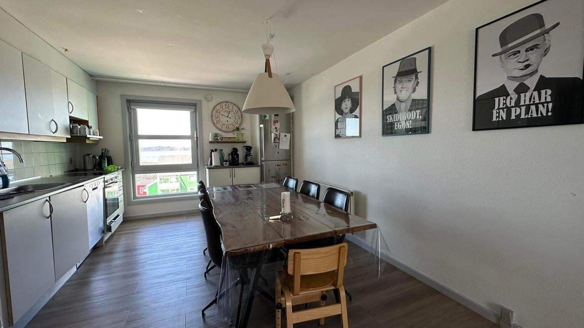 A bright kitchen features a long wooden dining table surrounded by several chairs, providing ample seating. Light floods in through a window, illuminating the modern cabinetry and appliances. Walls are adorned with framed black-and-white art, contributing to a contemporary aesthetic.