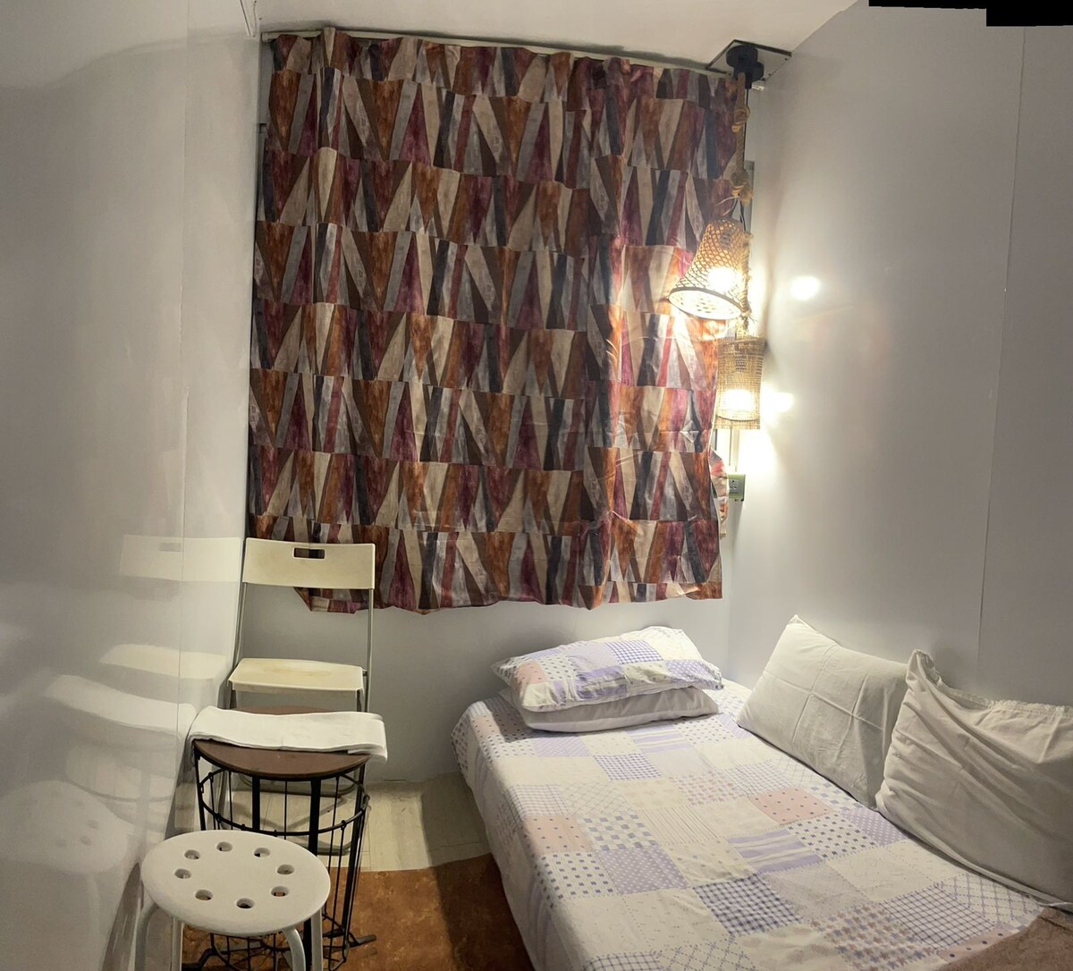 A compact sleeping area features a spacious bed with soft bedding, complemented by a colorful patterned curtain. A small white table and a wire stool provide seating options, while soft lighting emanates from decorative fixtures.