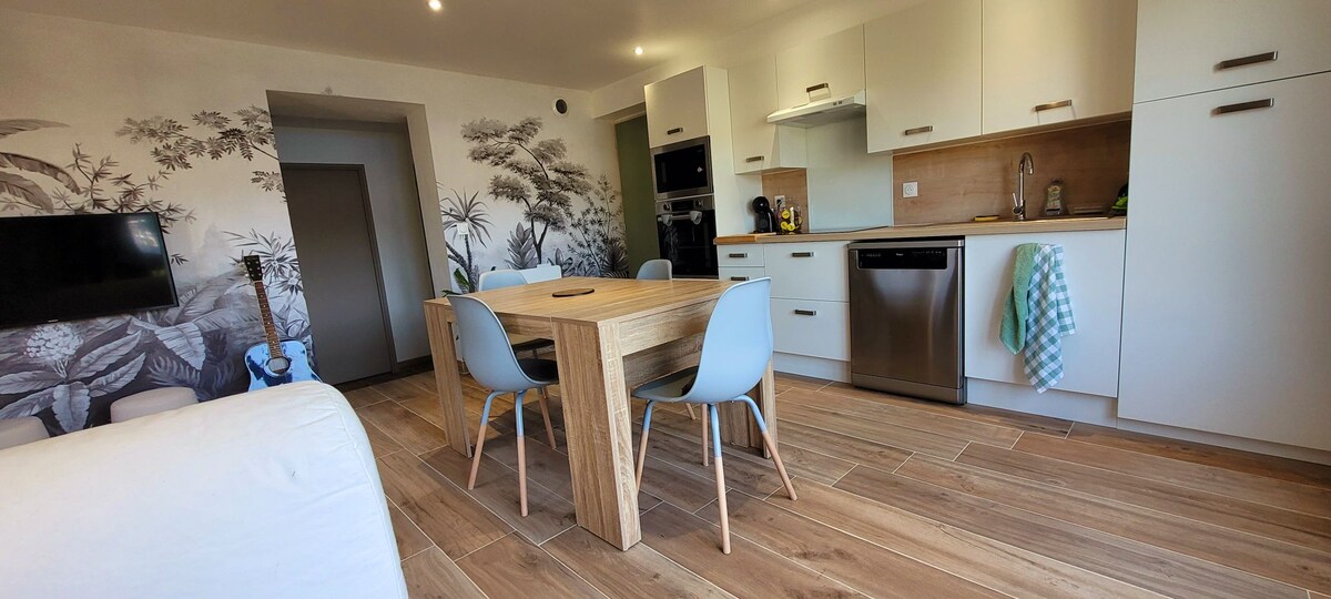 A modern kitchen and dining area features a wooden dining table surrounded by four light blue chairs. A sleek stainless steel dishwasher and oven are visible, alongside warm wood cabinetry. A large floral mural adorns one wall, complementing the space.