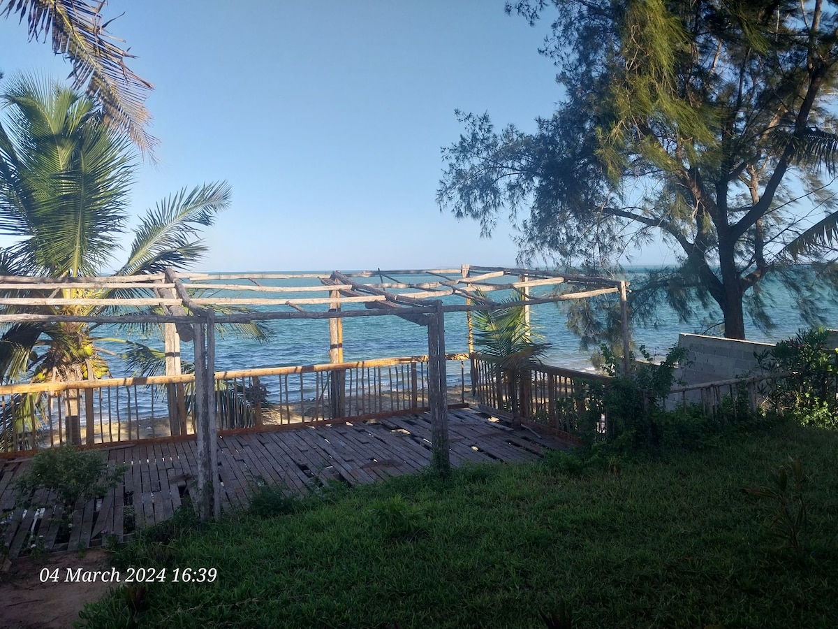 An open wooden deck is presented, bordered by green grass and palm trees, overlooking the ocean. The space is framed by a simple structure, allowing for unobstructed views of the water and coastline.