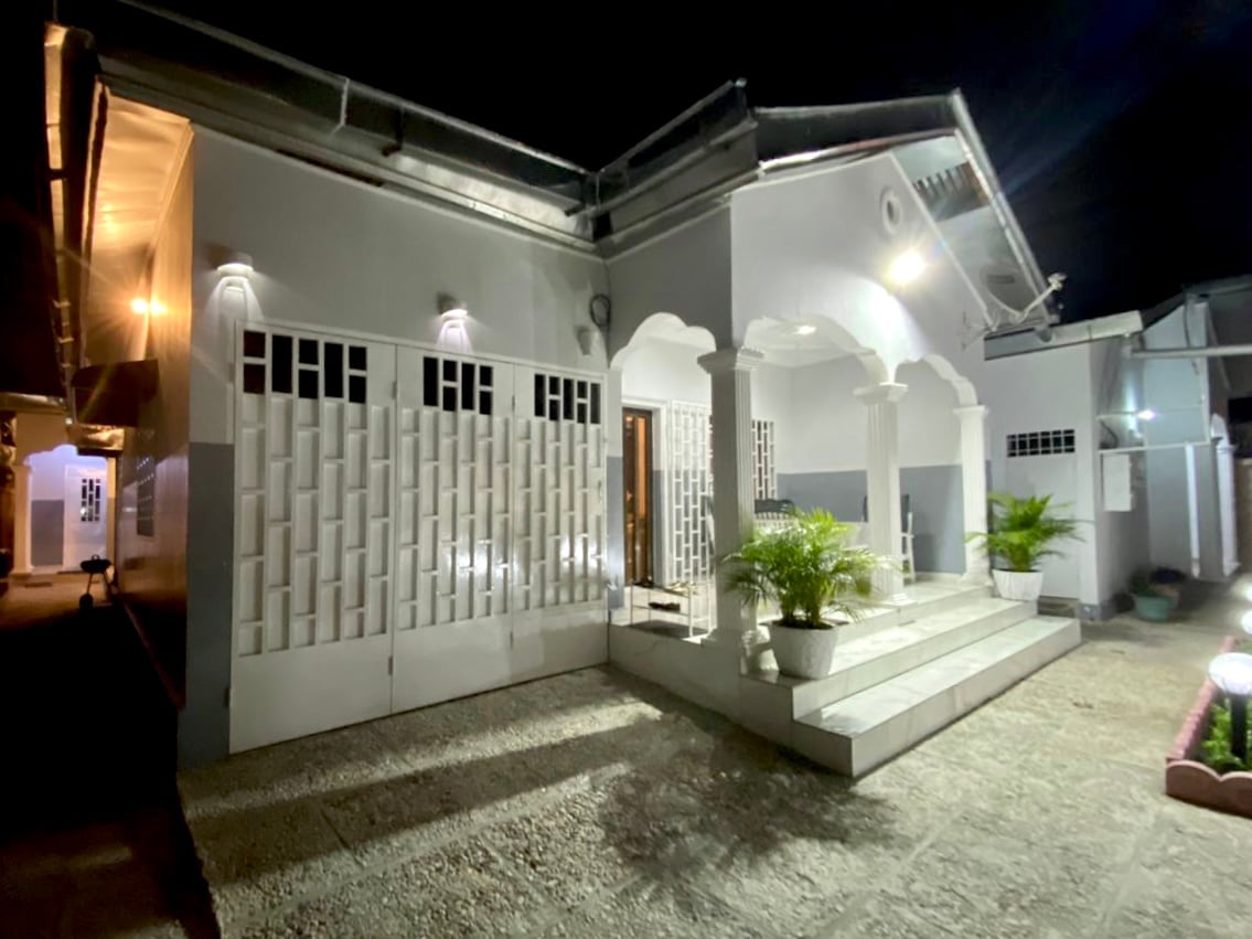 The exterior of the villa is presented at night, featuring a series of arched doorways and a staircase leading to the entrance. Soft lighting highlights the decorative facade and potted plants flank the steps, while a gravel pathway provides access from the side.