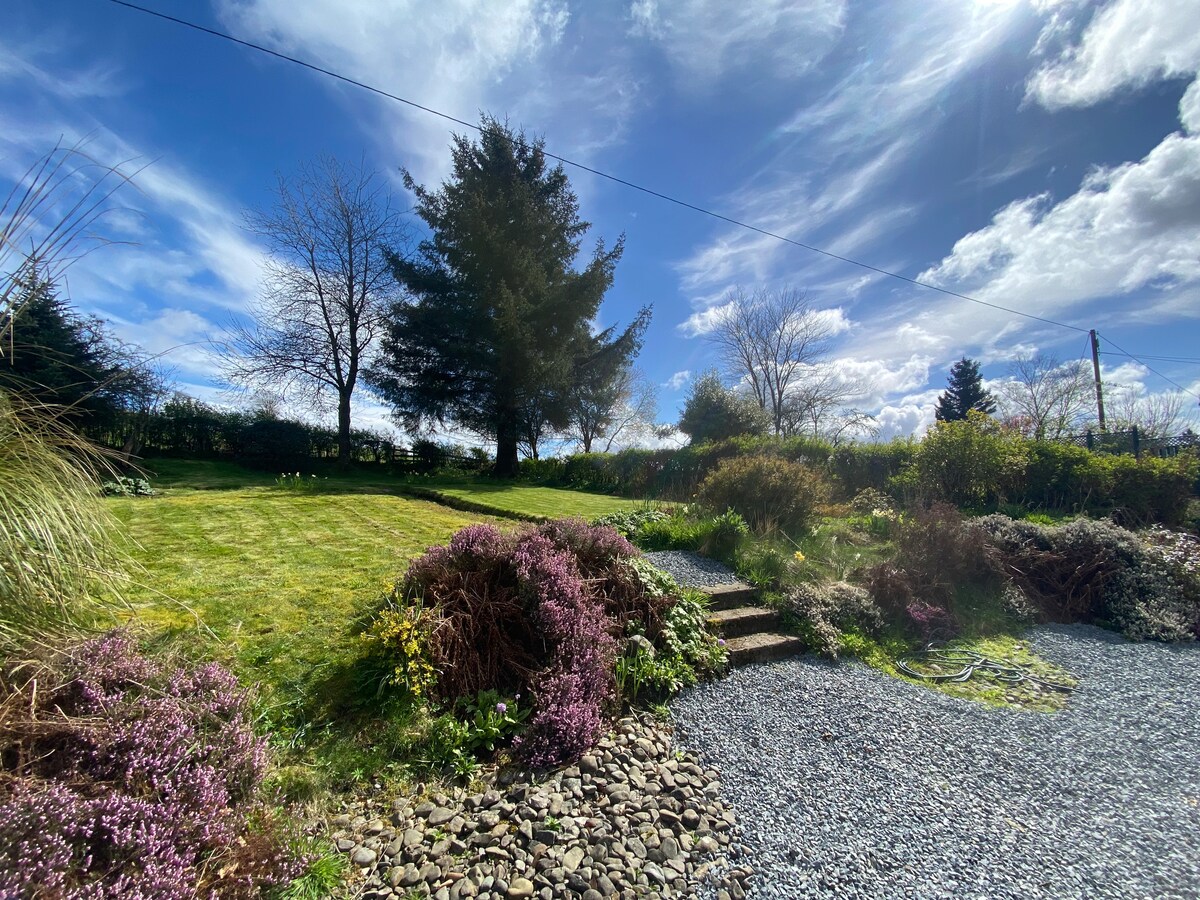 A spacious rear garden is shown, featuring a well-maintained lawn surrounded by mature trees and shrubs. Stone steps lead from a gravel pathway into the garden, with vibrant foliage and an expansive blue sky present in the backdrop.