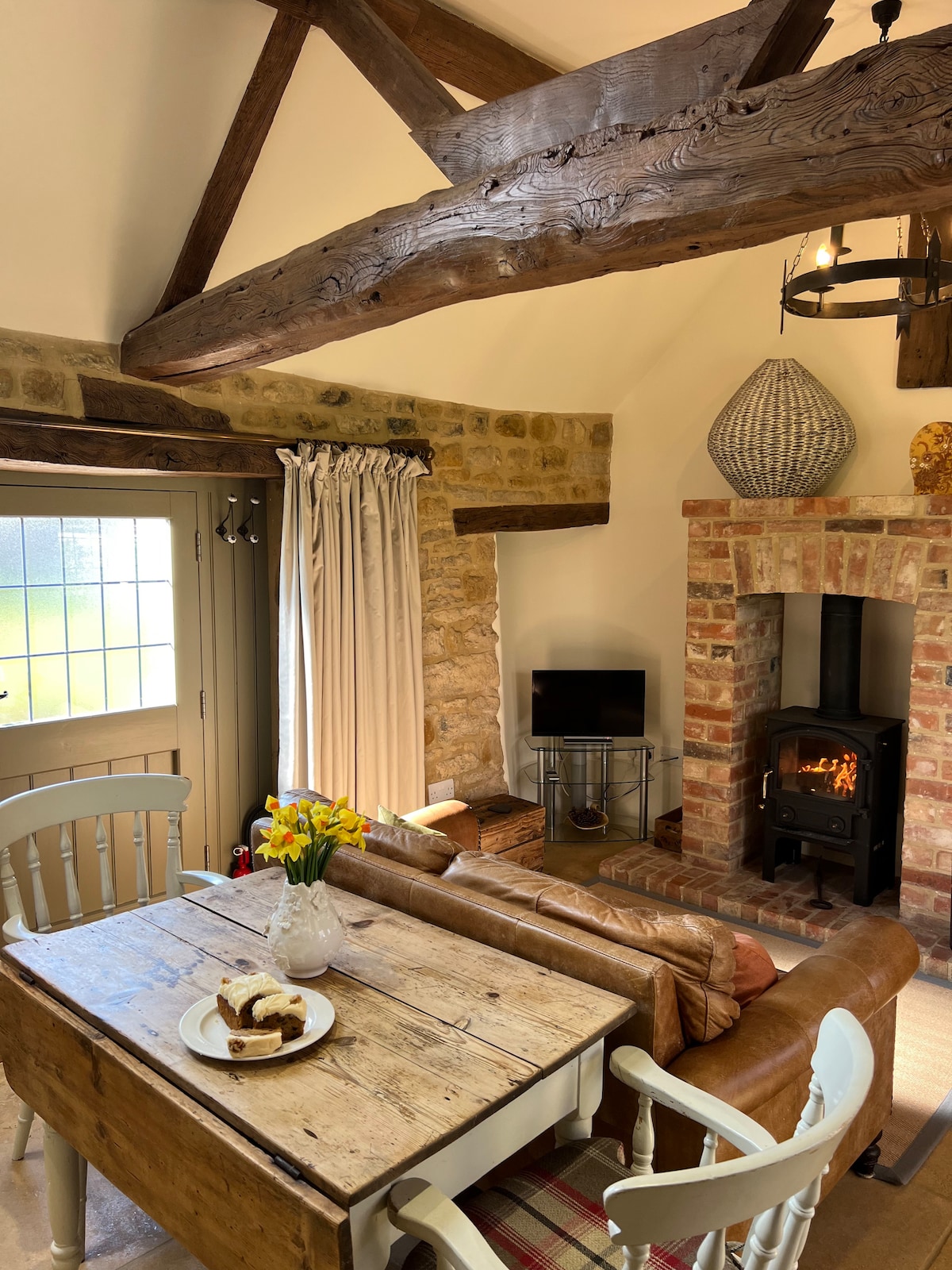 A bright and inviting open-plan space features exposed wooden beams and a cozy seating area. A rustic wooden dining table is set with a plate of cake and fresh flowers. A fireplace with a warm glow is complemented by a small television nearby.