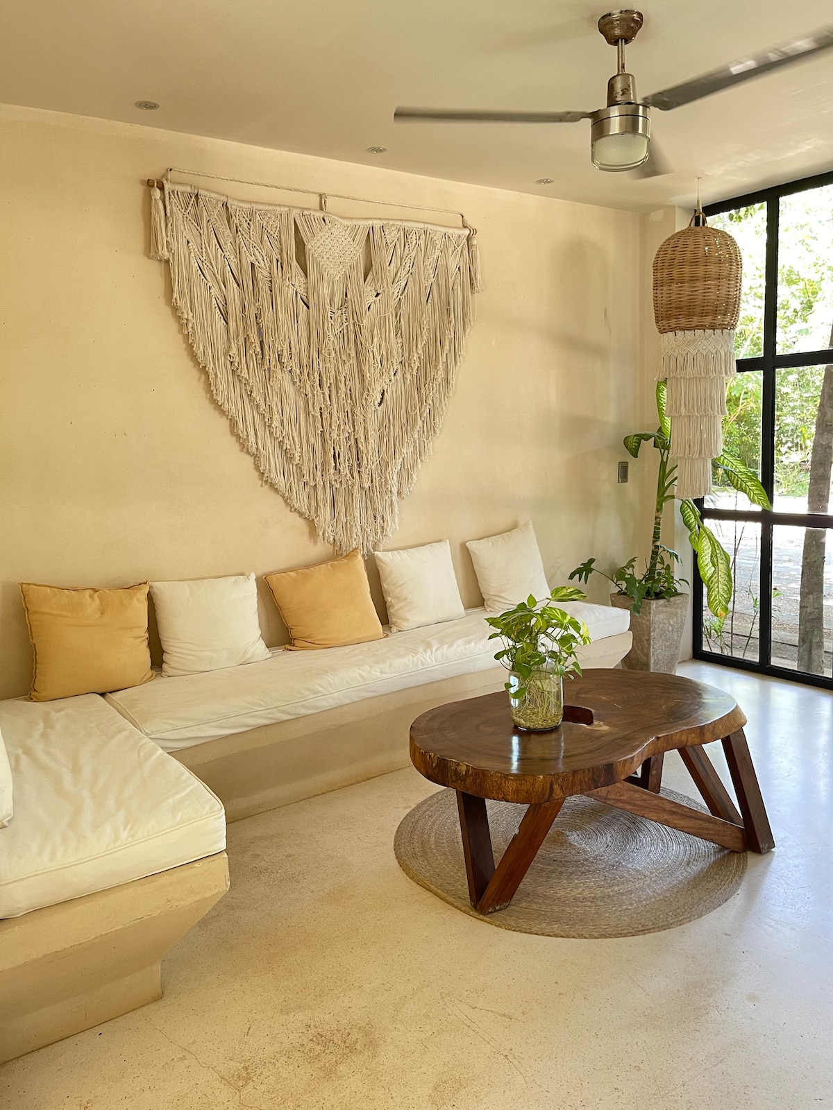 A minimalist living area features a spacious, L-shaped sofa adorned with neutral cushions. A wooden coffee table is centered on a round area rug, complemented by a small plant. An intricately woven wall hanging adds texture, while large windows allow natural light to flood the space.