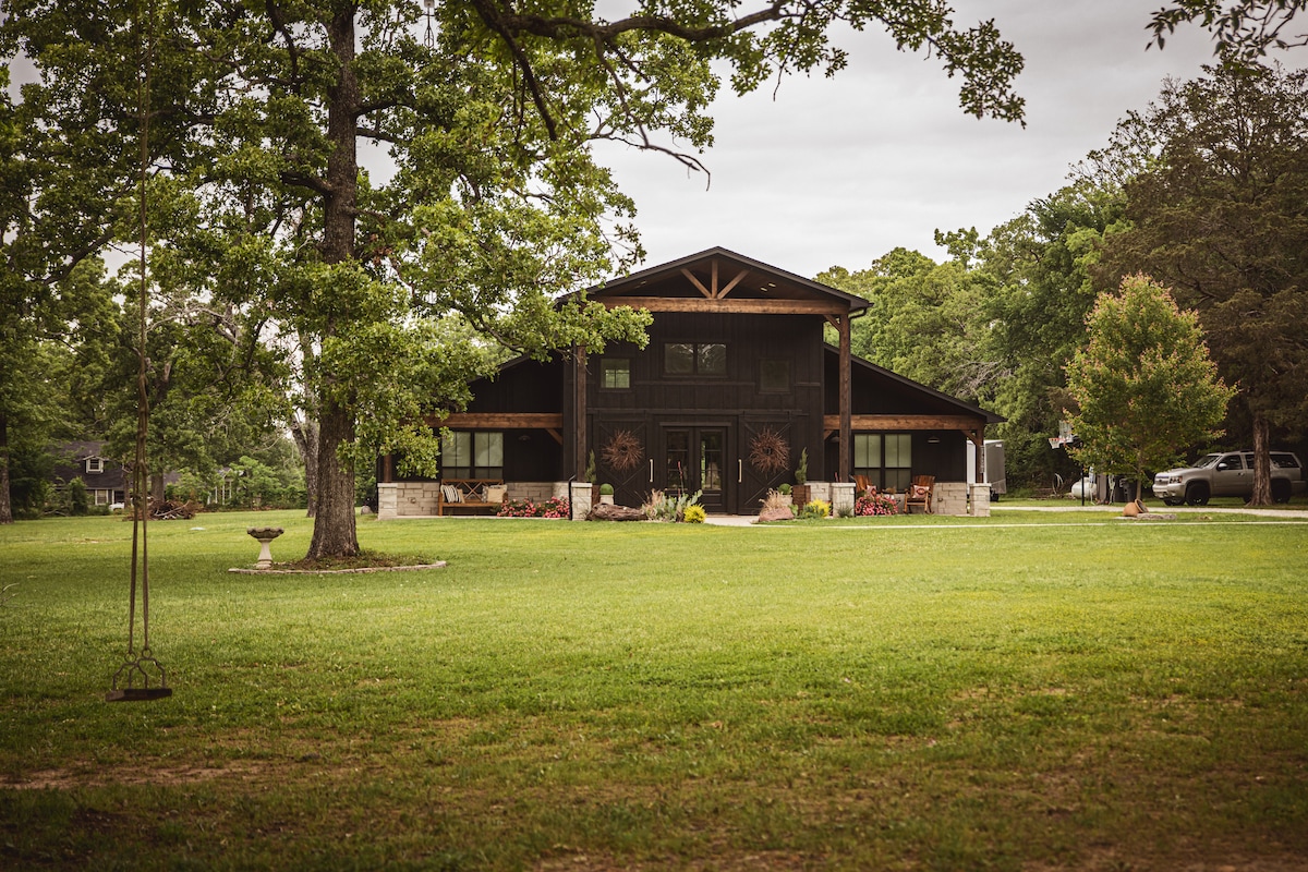 The barn house features a sturdy, weathered exterior with large windows framed by natural landscapes. A spacious lawn, accented by trees and colorful flowers, surrounds the structure, creating a serene outdoor space. The entrance is approached by a gravel path leading to the inviting front porch.