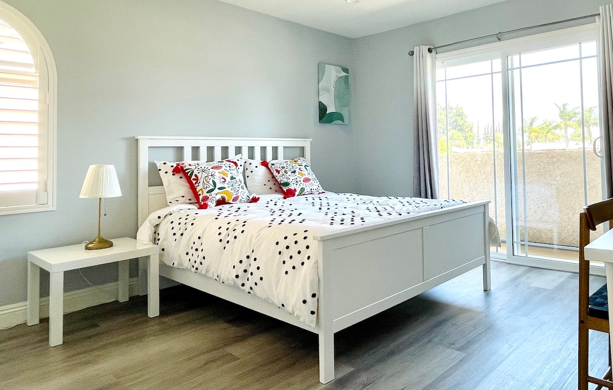 A bright bedroom features a spacious bed dressed with decorative pillows and a patterned quilt. A bedside table with a lamp is situated next to the bed. Large sliding doors provide natural light and lead to a balcony with views of greenery.