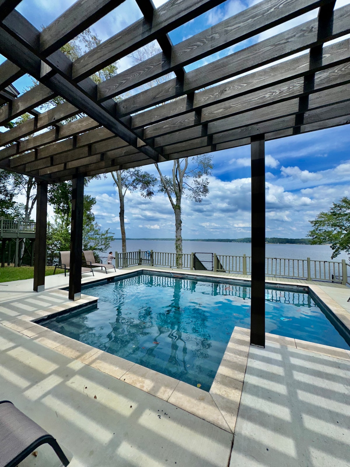 An outdoor pool is framed by a wooden pergola, allowing filtered sunlight to create a pattern on the water's surface. The surrounding area includes a view of the lake and trees, with seating options positioned nearby for relaxation.