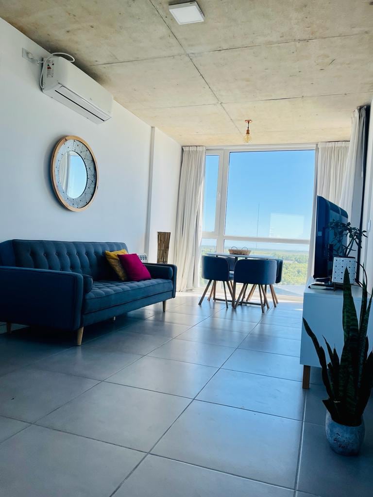 A bright living area features a modern blue sofa with colorful throw pillows. A round dining table with two chairs is positioned near a large window, allowing natural light to fill the space. A potted plant and minimalist decor items add a touch of greenery.