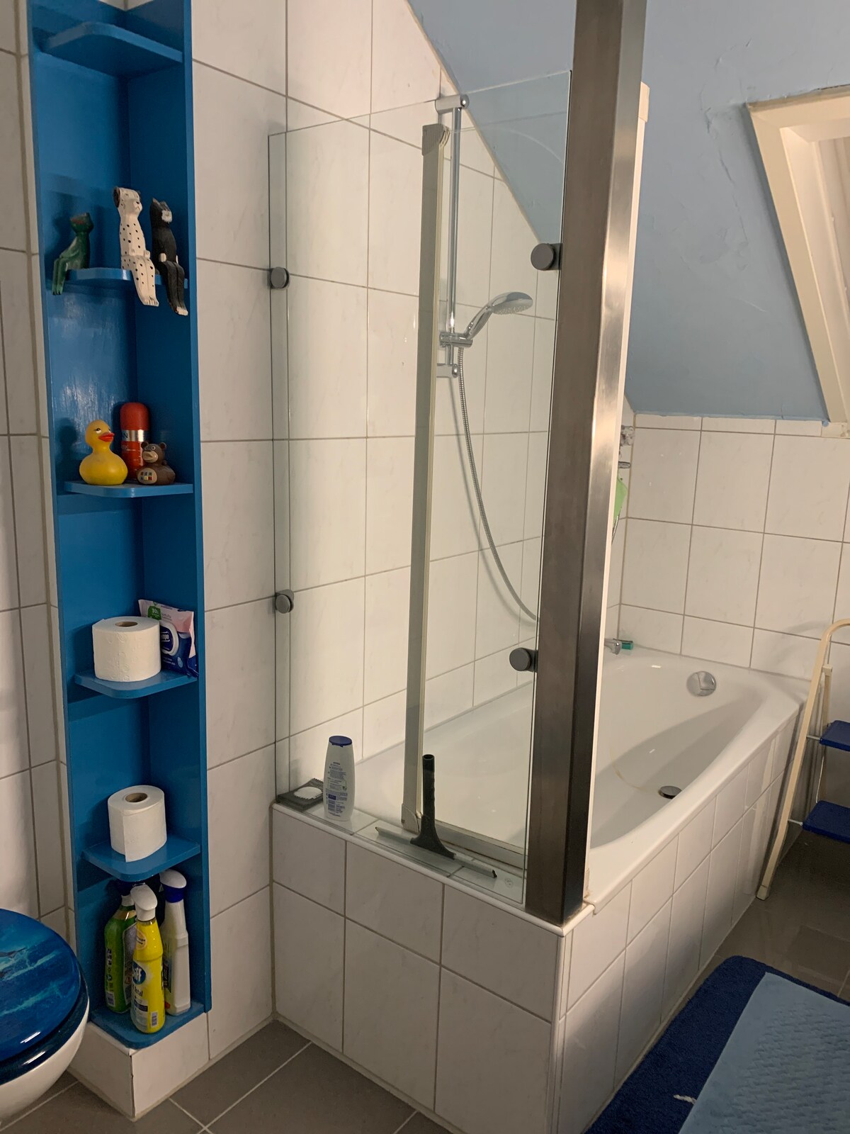 A modern bathroom features a glass shower enclosure adjacent to a bathtub. The tiled walls are complemented by a vibrant blue shelf displaying toiletries. A bath mat is positioned on the floor near the tub, and natural light filters through a small window above.