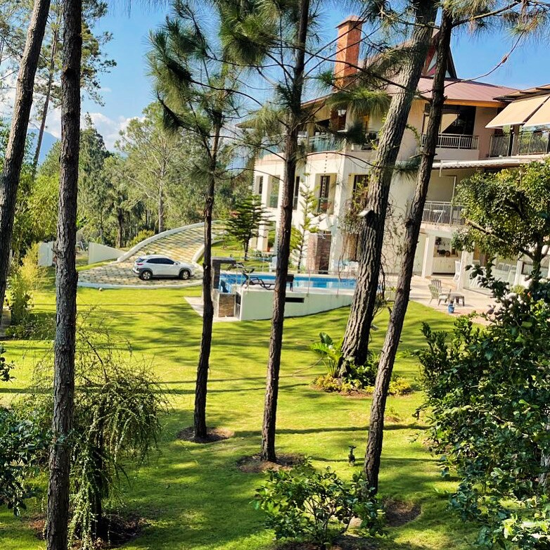 A private villa is surrounded by lush greenery and tall pine trees. A spacious lawn leads to a heated pool, visible behind the trees. The two-story structure features balconies with shaded areas, complemented by natural sunlight filtering through the foliage.