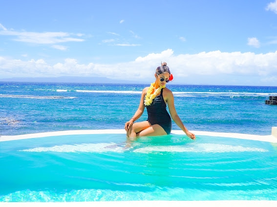 A person relaxes in a private pool with ocean views. The bright blue water contrasts against the horizon, and gentle waves can be seen in the distance. The sky is clear with a few clouds, enhancing the tranquil outdoor atmosphere.