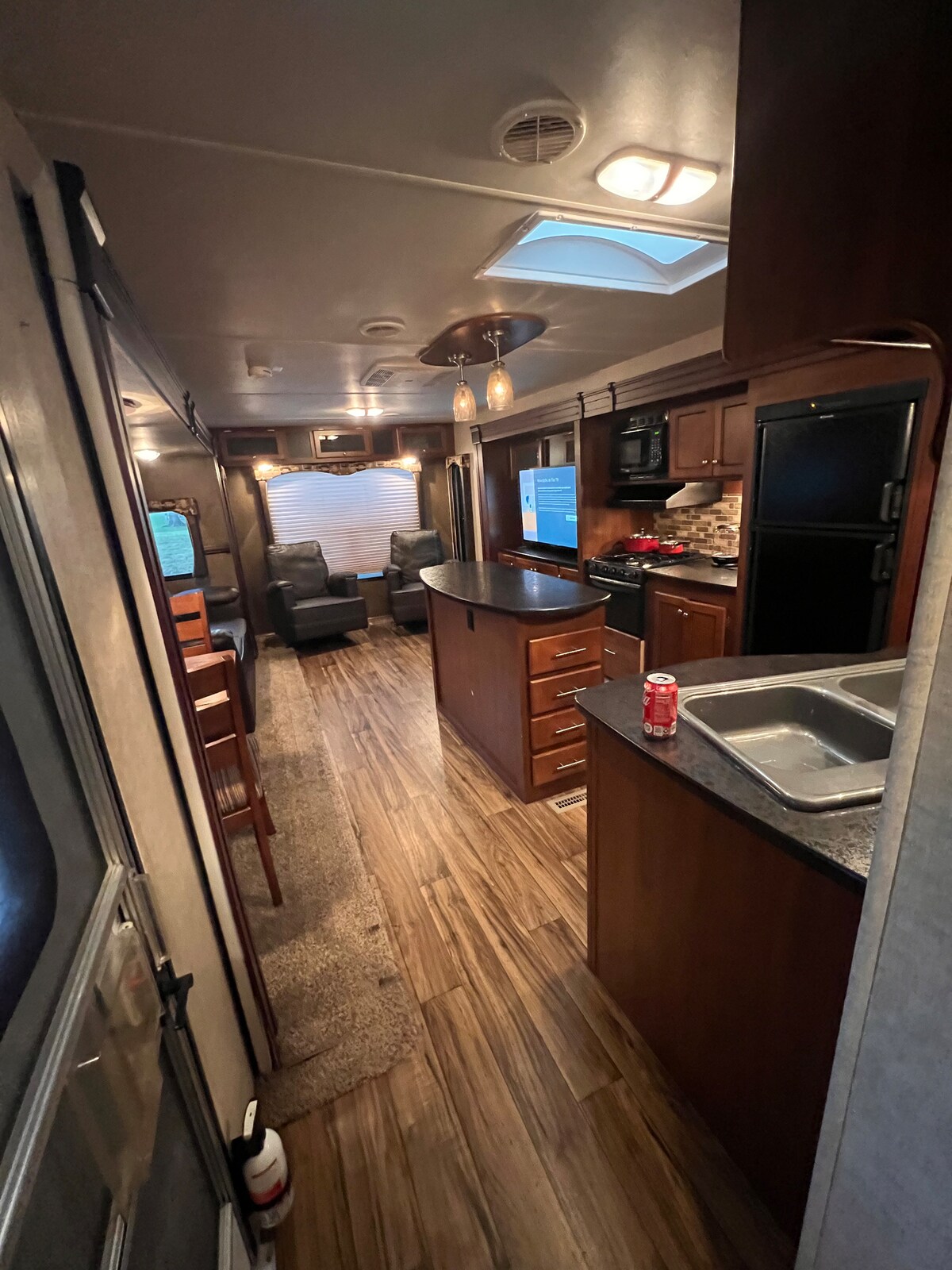 An interior view of the RV is displayed, showcasing a cozy layout with wooden cabinets and countertops. A minifridge and microwave are visible. The carpeted area extends to seating, with a small table and chairs positioned nearby. Skylights provide natural light to the space.