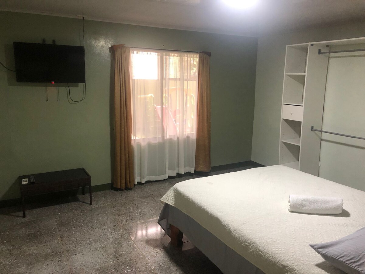 A spacious room features light green walls and a large window adorned with sheer curtains. A comfortable bed is neatly made with a white blanket and a towel placed on top. A flat-screen TV is mounted on the wall, and a simple wooden table is positioned nearby.