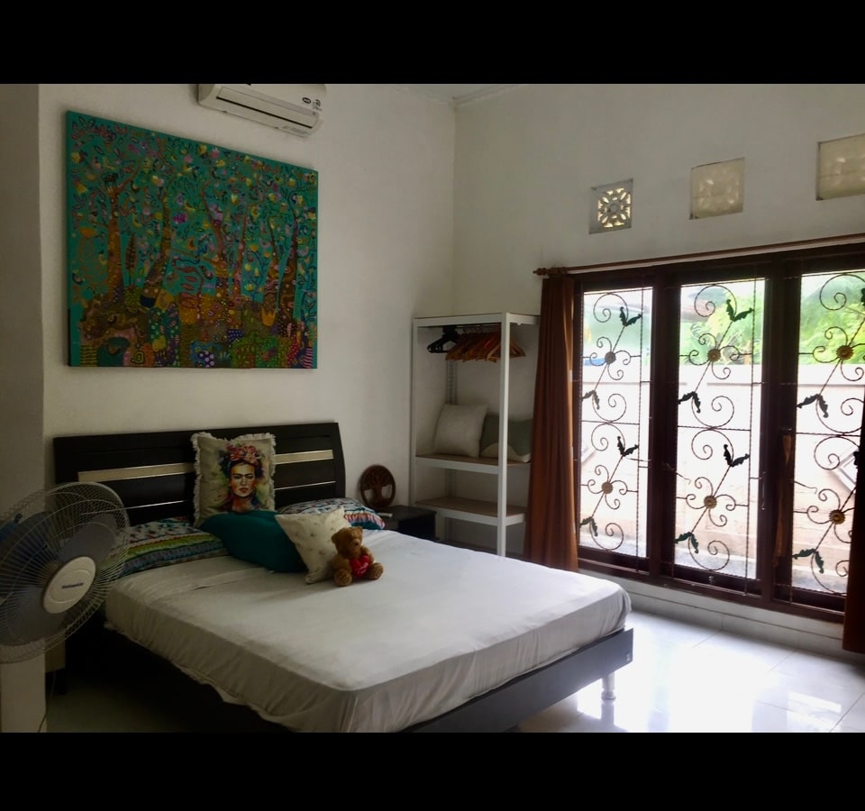 A bright bedroom features a spacious bed with a colorful decorative blanket. A large piece of wall art depicting trees adds a touch of vibrancy. Natural light enters through the ornate windows, complemented by a fan and a shelf with various items thoughtfully arranged.