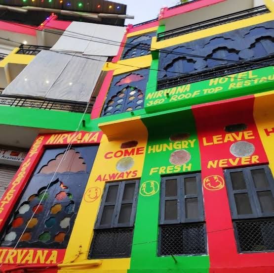 A vibrant multi-colored building displays an energetic façade decorated with traditional arches and large windows. The text 'NIRVANA HOTEL' and additional messages are prominently featured, contributing to the lively atmosphere. It is situated in a bustling area, indicating proximity to various shops and restaurants.