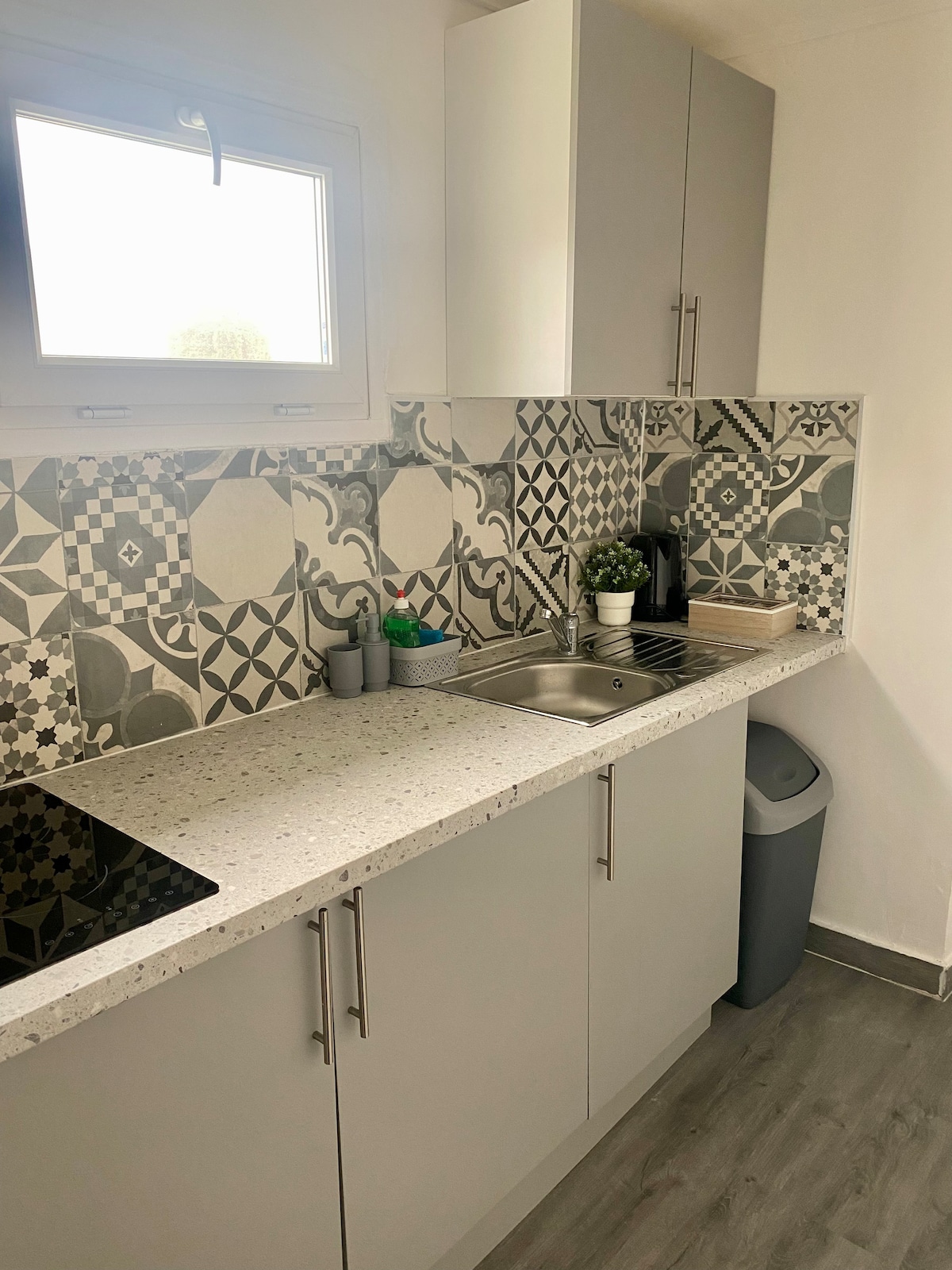 The kitchenette features modern grey cabinetry and intricate patterned tiles on the backsplash. A stainless steel sink and an electric cooktop are included, along with a small counter space for meal preparation. Natural light enters through a window above the sink.