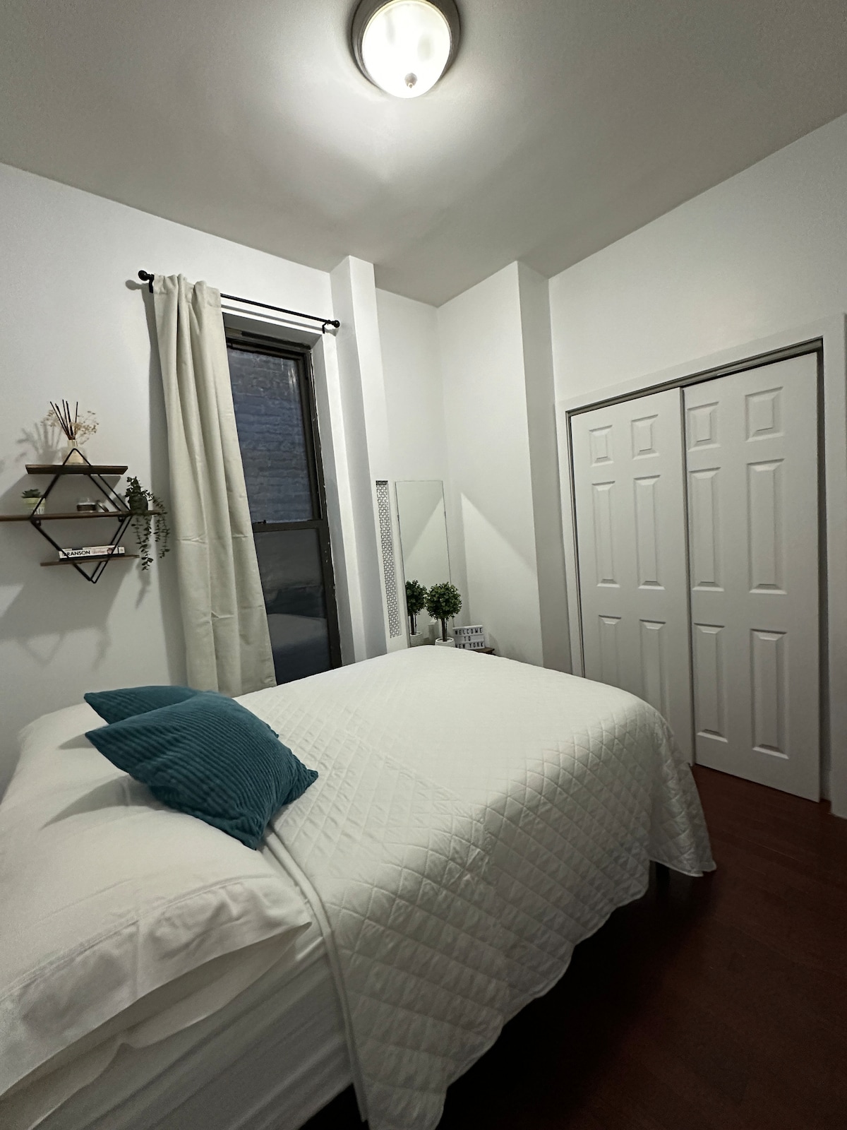 A well-lit bedroom features a spacious bed adorned with a light quilt and a decorative pillow. A full-length mirror is positioned beside the bed, reflecting the room's bright atmosphere. Two closets with sliding doors offer storage space, while a small plant adds a touch of greenery.