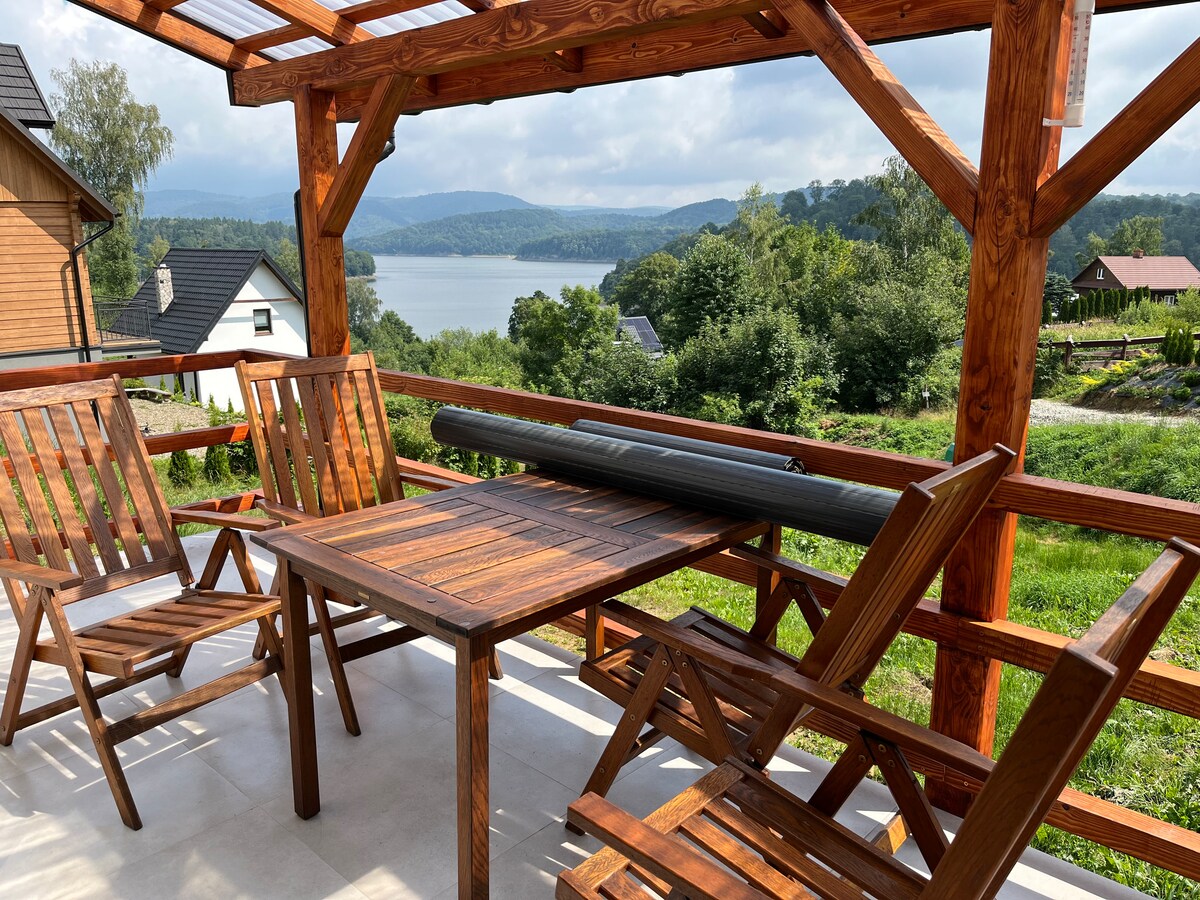 A wooden terrace features a table with four matching chairs, providing a scenic view of the lake and surrounding greenery. The outdoor space is shaded by a wooden pergola, with soft clouds visible in the sky above.