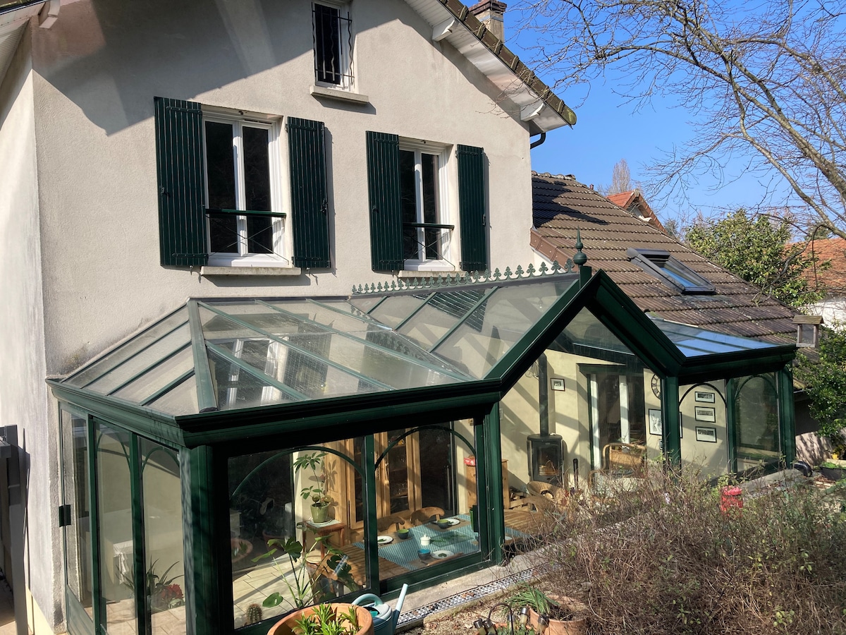The image showcases a stylish glass veranda attached to a house, featuring a green frame and multiple panels reflecting sunlight. The exterior reveals large windows and a tiled roof, surrounded by a small garden area with various plants, creating a harmonious connection between indoors and outdoors.