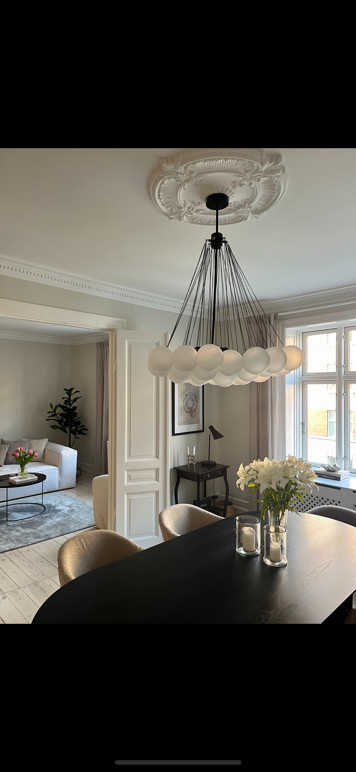 An elegant dining area features a dark table paired with soft, round chairs. Above, a striking chandelier with white globes creates ambient light, while a vase of flowers adds a touch of freshness. A glimpse of the adjoining living area is visible through an open door.