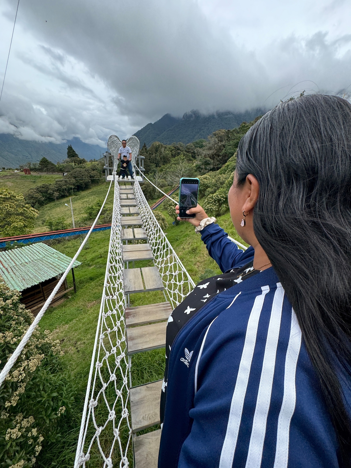 A suspension bridge spans a scenic landscape, connecting two areas of greenery. A person captures a photo of two people seated in front of a winged sculpture. Dramatic clouds form a backdrop over the distant mountains, enhancing the natural beauty of the setting.