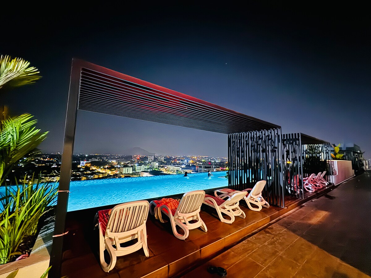 A rooftop infinity pool offers panoramic city views under the night sky. White lounge chairs line the wooden deck, with a stylish partition providing privacy. Soft lighting enhances the serene atmosphere, inviting relaxation and enjoyment of the surrounding scenery.