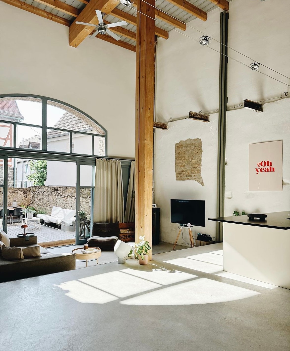 A spacious living area is characterized by high ceilings and large windows allowing natural light to fill the room. Wooden beams and a light-colored wall complement the minimalist decor. A cozy seating area and a TV are visible in the background, with outdoor seating accessible through the glass doors.