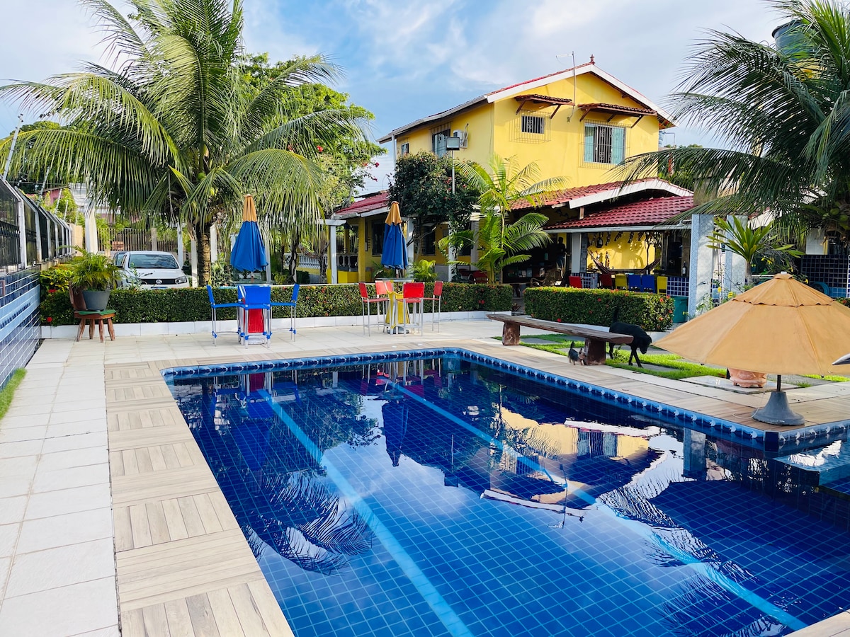 An outdoor swimming pool is surrounded by palm trees, sun loungers, and a shaded area with umbrellas. The pool reflects the sky, and colorful furniture is positioned nearby, creating a relaxing atmosphere. A well-maintained garden adds greenery to the space.
