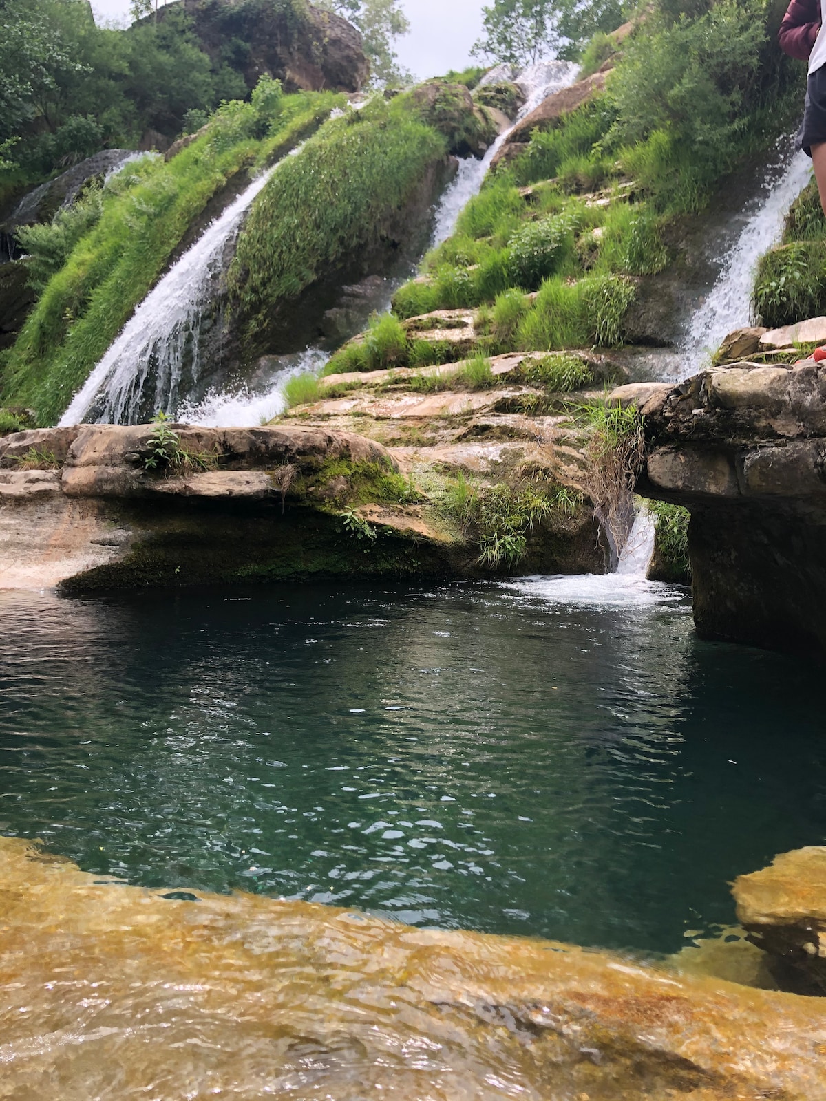 A natural waterfall cascades over rocky terrain, creating gentle streams that flow into a clear, still pool. Lush greenery surrounds the area, adding a vibrant touch. The rugged rocks form a varied texture alongside the water, inviting exploration and relaxation in this serene outdoor setting.