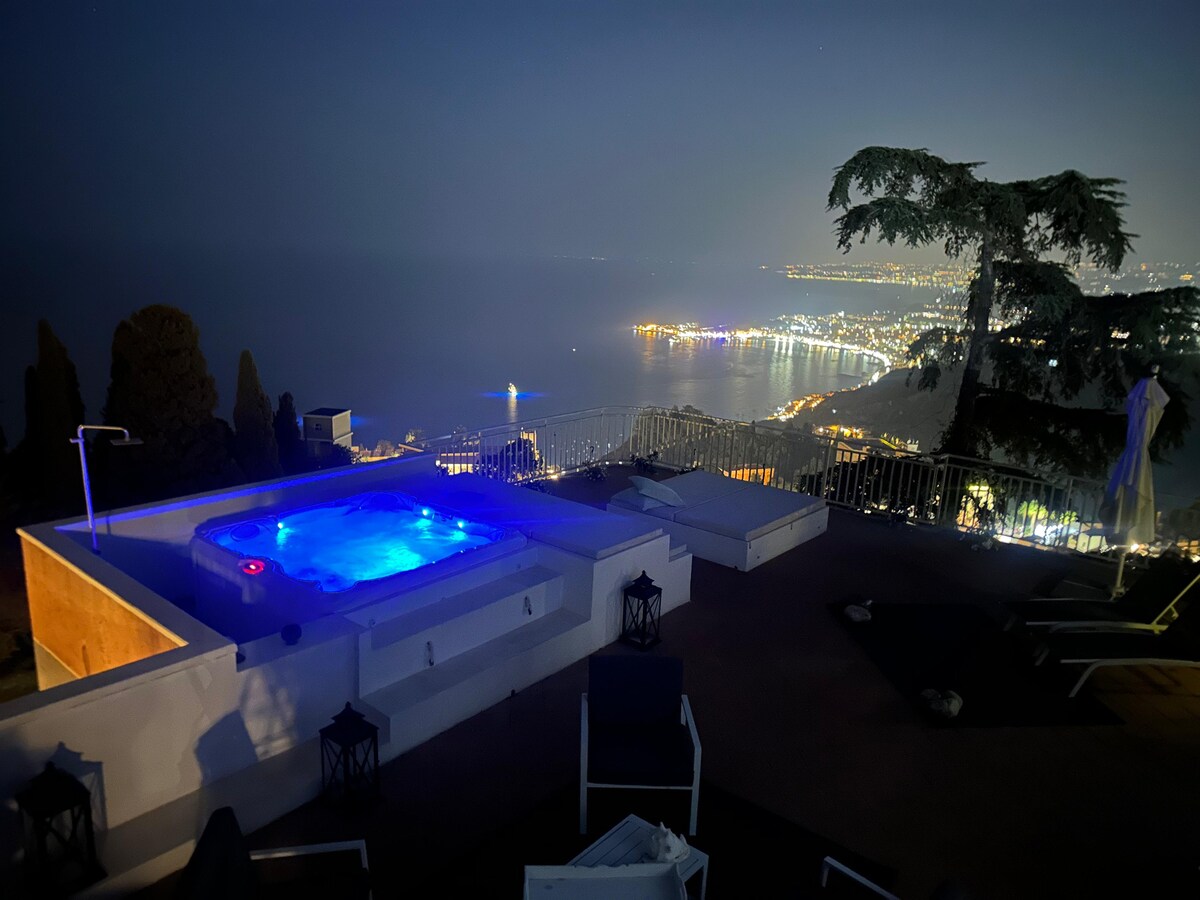 A private terrace is illuminated by the glowing blue lights of a jacuzzi, overlooking a scenic night view of the coastline. Soft lounge chairs and decorative lanterns create a relaxing environment, framed by trees against a backdrop of a starry sky.