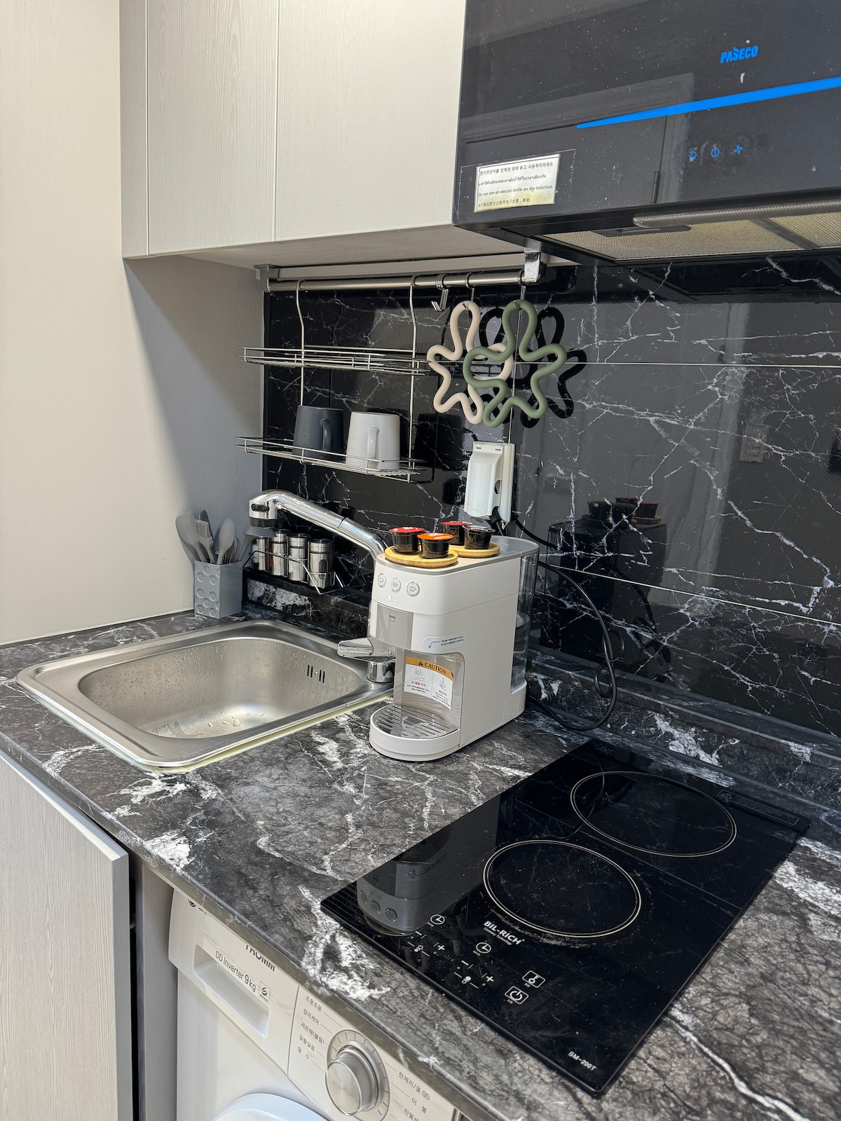 A modern kitchen space features a sleek marble countertop with an integrated sink. A coffee machine and a minimalist stovetop are positioned nearby, while various utensils and containers are organized neatly on the shelves above, providing a functional cooking area.