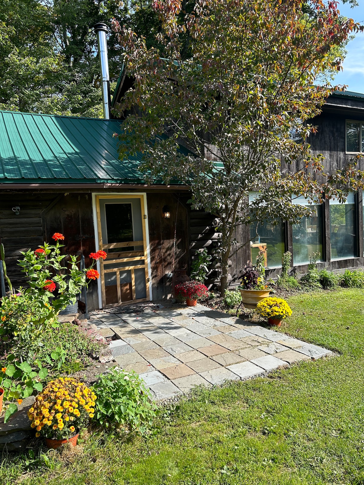 An inviting entryway is framed by a stone pathway leading to a wooden door. Brightly colored flower pots with seasonal blooms, including red and yellow flowers, are positioned along the pathway, while a lush green lawn offers a natural backdrop.